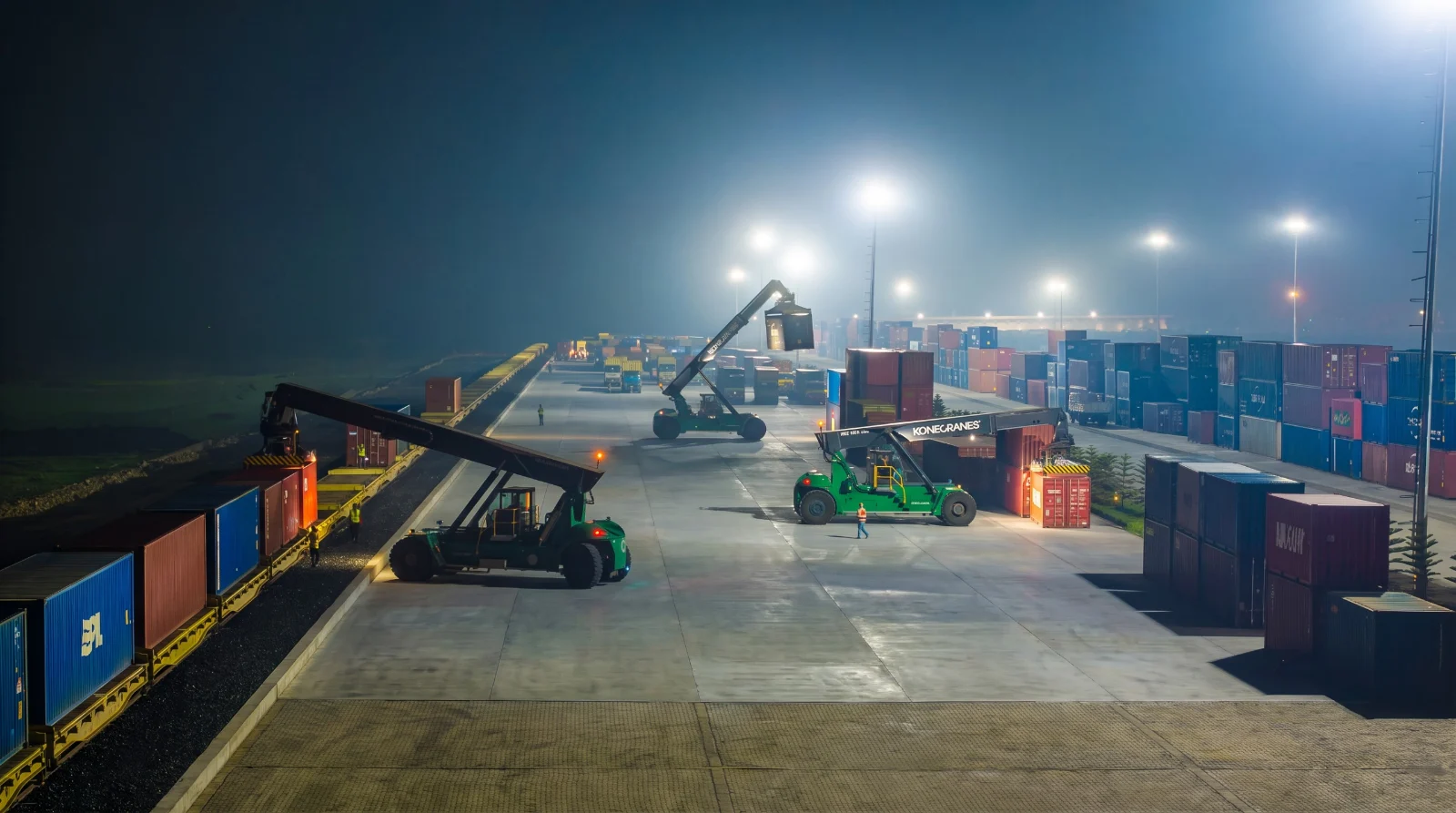 A dry port at night: reach stackers working container stacks under floodlights, with a loaded train alongside