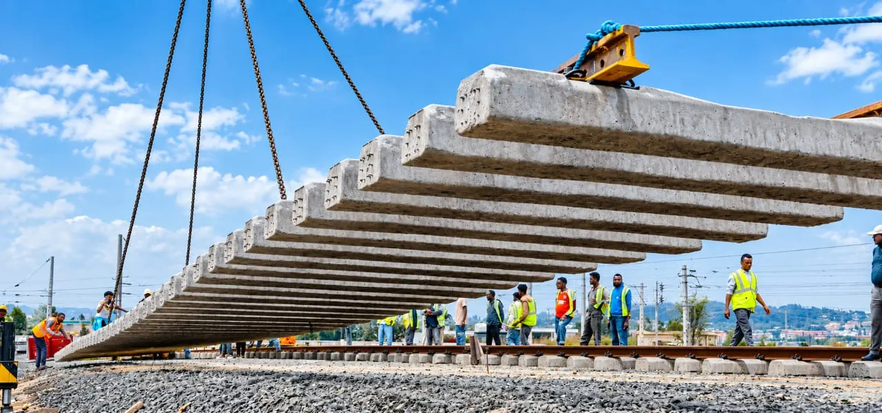 A crane lowering a bundle of concrete sleepers over a new track while a line of workers in high-visibility vests looks on