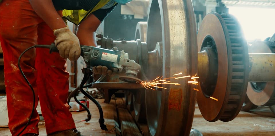 A technician in orange overalls and gloves working an angle grinder against a railway wheelset, sparks arcing off the wheel