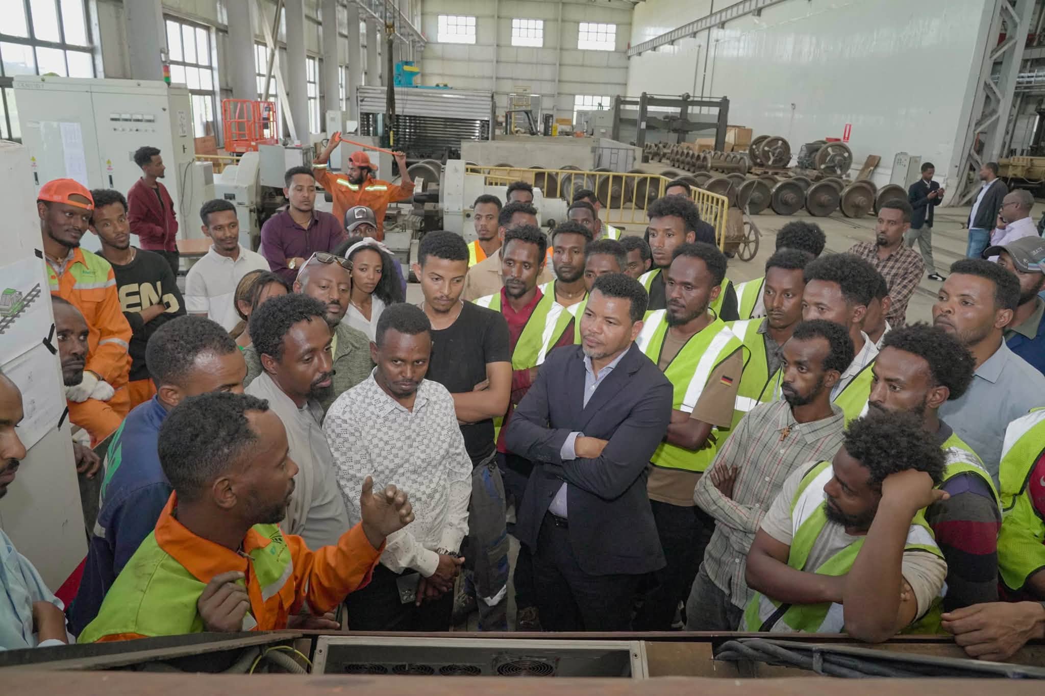 A large group of EDR workshop staff in high-visibility vests and overalls listening to a colleague inside a maintenance hall, rows of wheelsets stacked behind them